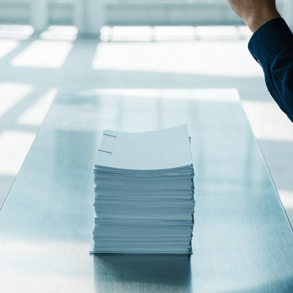 A neat stack of blank stapled procedure pages resting on a steel bench in cool daylight, a hand leaving frame at the top